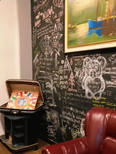 Pared-pizarra de la sala de Xantico cubierta de mensajes y dibujos en tiza, con un cuadro enmarcado, un juego de mesa en una maleta y un sillón de cuero rojo.