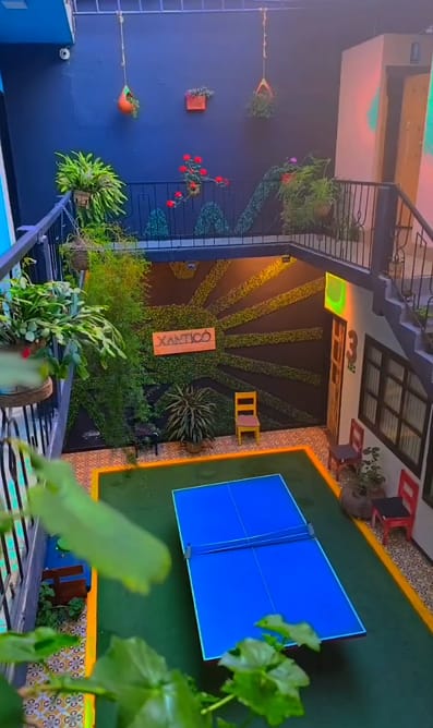 El patio de Xantico Hostal visto desde un balcón: mesa de ping-pong azul, muro oscuro con rayos de follaje verde y el letrero Xantico, plantas colgantes y balcones.