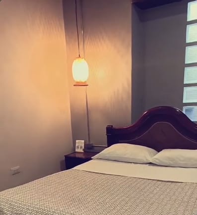 Habitación doble de Xantico: cama doble con cabecero curvo de madera oscura, dos almohadas blancas, colcha estampada gris y una lámpara colgante de luz cálida junto a la mesa de noche.