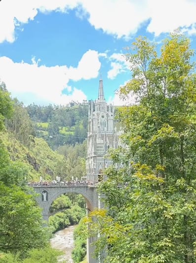 El santuario de Las Lajas asoma entre árboles verdes, con el puente y el río abajo, bajo un cielo azul con nubes blancas.