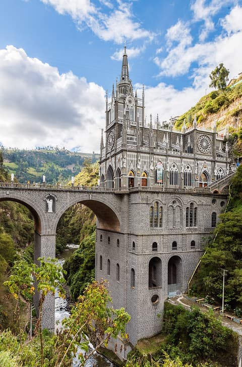 Santuario de Las Lajas: iglesia neogótica de piedra gris sobre un puente de arcos en el cañón del río Guáitara, rodeada de vegetación y cielo con nubes.