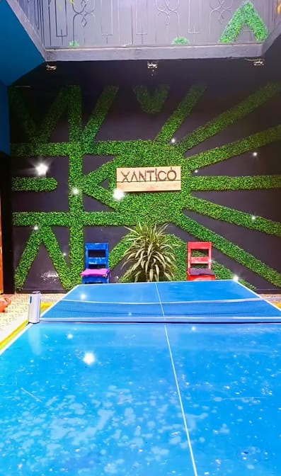 Mesa de ping-pong azul frente al muro de rayos verdes con el letrero Xantico, una silla azul y otra roja.