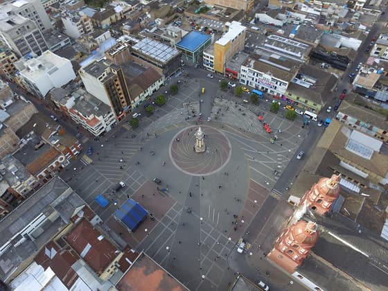 Vista aérea de la Plaza 20 de Julio en el centro de Ipiales: una plaza amplia con un monumento circular en el centro, rodeada de edificios y las cúpulas de ladrillo de una iglesia en la esquina.