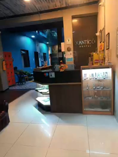 Recepción con mostrador oscuro y letrero Xantico Hostal; al fondo, un pasillo de paredes azules y lockers naranjas.